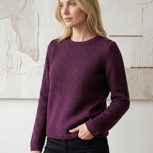 May include: A long-sleeved, crew-neck sweater in a deep purple hue. The knitwear has a textured appearance and is paired with black pants. The model is standing in front of a neutral-toned, abstract background.