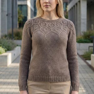 May include: A brown sweater with a crew neck and long sleeves. The sweater features a detailed leaf pattern across the upper chest. The model is wearing tan pants. The background shows a modern building.