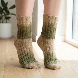 May include: A pair of ribbed knit socks with a color gradient. The socks transition from a light tan at the foot to a green hue at the mid-calf. The socks are worn on a person's feet, with the legs visible from the ankles down.
