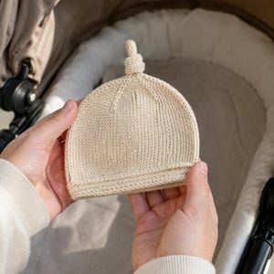 May include: A cream-coloured knitted baby hat with a knotted detail at the crown. The hat is held in front of a pram. The hat is made of a soft, textured yarn. The hat is a neutral colour, suitable for a baby.
