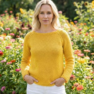 May include: A woman wearing a mustard yellow knit sweater with long sleeves and a boat neck. The sweater is paired with white pants. The background features a colorful flower garden.