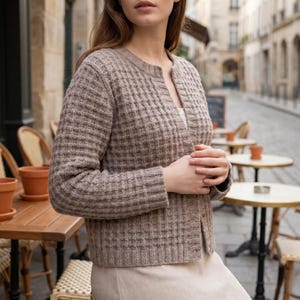May include: A light brown, open-front cardigan with a textured, grid-like pattern. The long-sleeved sweater is worn over a white top and a beige skirt. The background shows a European street scene with outdoor cafe tables and chairs.