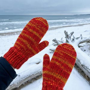 May include: A pair of red and yellow striped knitted mittens. The mittens appear thick and warm, suitable for cold weather. The background shows a snowy beach and the sea.