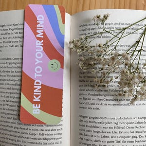 Könnte beinhalten: Ein farbenfroher Lesezeichen mit dem Text "BE KIND TO YOUR MIND" in Weiß. Das Design zeigt wellenförmige Streifen in Rosa-, Orange-, Blau- und Grüntönen mit einem lächelnden Gesichts-Symbol. Das Lesezeichen liegt in einem offenen Buch.