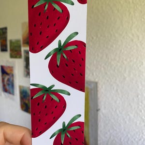 Könnte beinhalten: Ein weißes Lesezeichen mit einem sich wiederholenden Muster aus großen, roten Erdbeeren mit grünen Blättern. Die Erdbeeren haben kleine schwarze Samen. Das Lesezeichen wird von einer Hand gehalten.