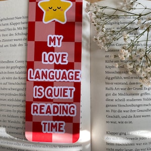 Könnte beinhalten: Lesezeichen mit rot-weißem Karomuster und dem Text "MY LOVE LANGUAGE IS QUIET READING TIME" sowie einem gelben Stern. Das Lesezeichen liegt auf einem offenen Buch.