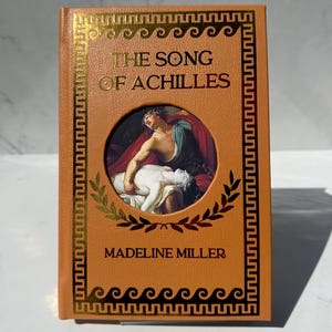May include: A hardcover book titled "THE SONG OF ACHILLES" by Madeline Miller. The cover is a warm orange with gold accents, including a decorative border and the title's text. A circular image depicts a classical painting.