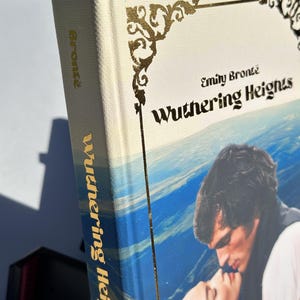 May include: A hardcover book titled "Wuthering Heights" by Emily Bront&euml;. The book has a cream-colored cover with gold decorative accents and text. The spine and cover feature the title in gold lettering. The cover art depicts a romantic scene with two people kissing.
