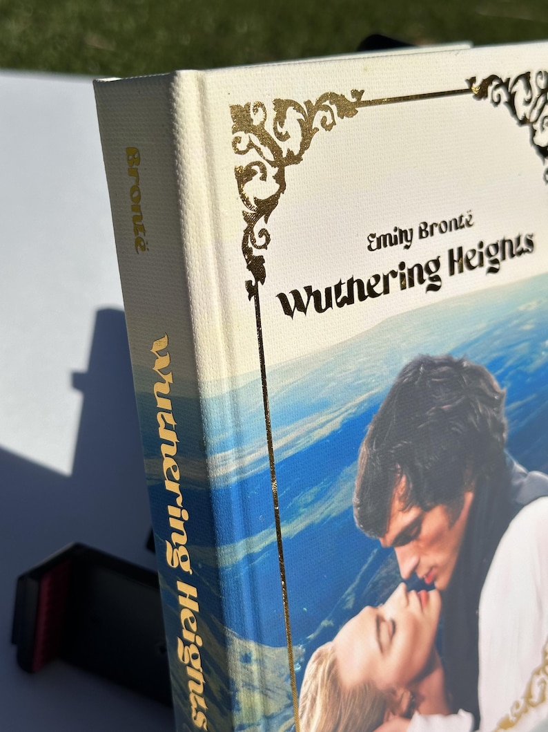 May include: A hardcover book titled "Wuthering Heights" by Emily Bront&euml;. The book has a cream-colored cover with gold decorative accents and text. The spine and cover feature the title in gold lettering. The cover art depicts a romantic scene with two people kissing.