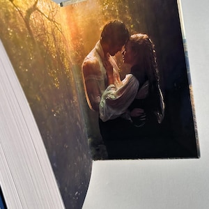 May include: An open book with a romantic image of a couple embracing in a forest setting. The image features a man and woman in a tender moment, with a soft, golden light filtering through the trees. The book's pages are visible, with a white and blue spine.