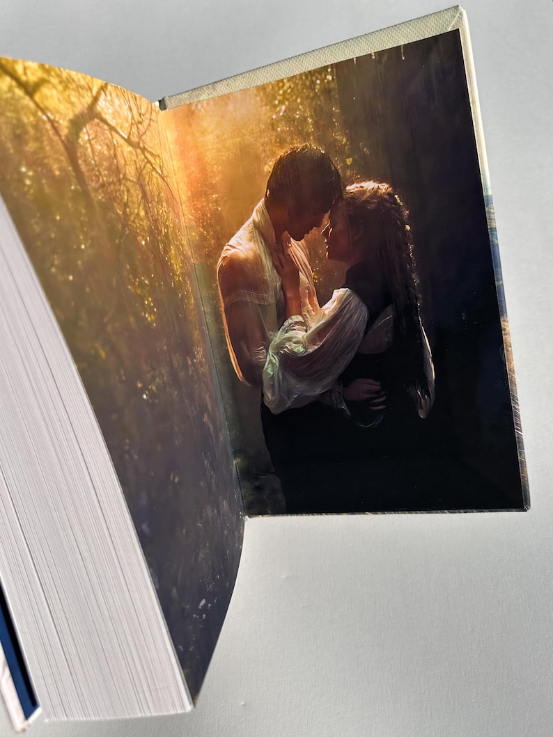 May include: An open book with a romantic image of a couple embracing in a forest setting. The image features a man and woman in a tender moment, with a soft, golden light filtering through the trees. The book's pages are visible, with a white and blue spine.