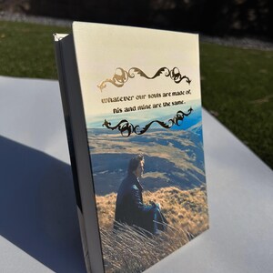 May include: A hardcover book with a landscape image and the quote, "whatever our souls are made of, his and mine are the same." The cover features a man sitting in tall grass with a mountain backdrop. Gold filigree accents the text.