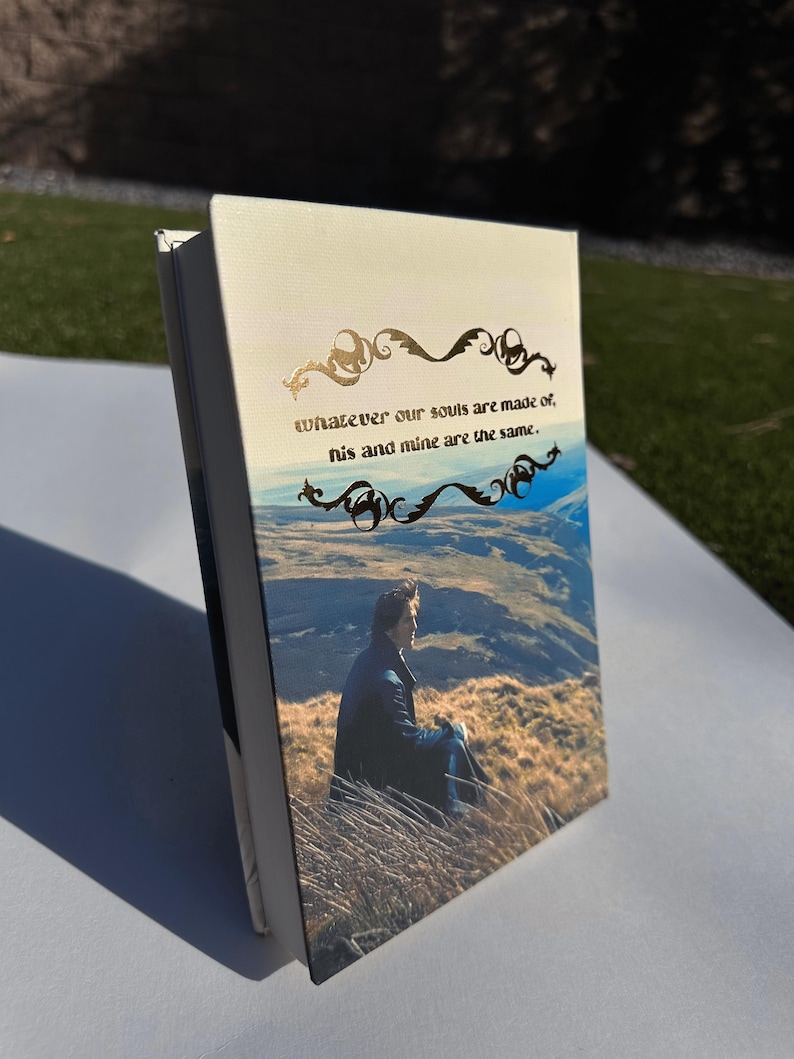 May include: A hardcover book with a landscape image and the quote, "whatever our souls are made of, his and mine are the same." The cover features a man sitting in tall grass with a mountain backdrop. Gold filigree accents the text.