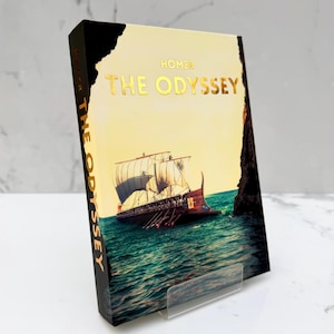 May include: A hardcover book titled The Odyssey by Homer, featuring a gold-foiled title on a cream-colored cover. The cover art depicts an ancient wooden ship with sails on a blue sea near a dark cliff. The spine also displays the title in gold.