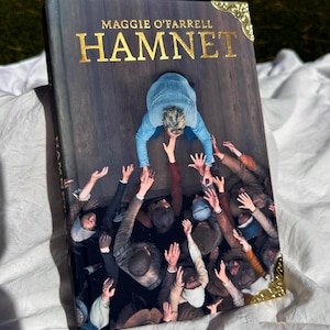May include: A hardcover book titled "HAMNET" by Maggie O'Farrell. The cover features a person in a blue shirt reaching down to many hands reaching up. Gold corner protectors and text on a dark background.