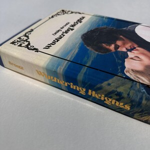 May include: A hardcover book titled "Wuthering Heights" by Emily Bront&euml;. The cover features a romantic image of two people kissing against a backdrop of a blue sky and rolling hills. The book's spine displays the title in gold lettering.