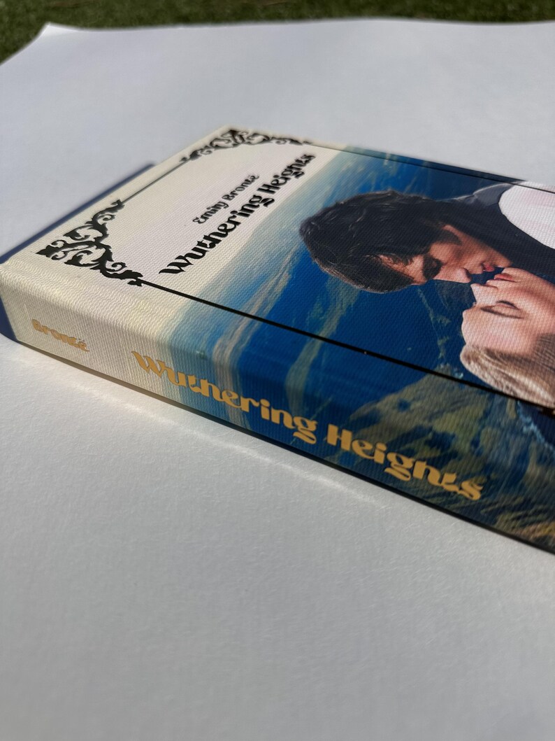 May include: A hardcover book titled "Wuthering Heights" by Emily Bront&euml;. The cover features a romantic image of two people kissing against a backdrop of a blue sky and rolling hills. The book's spine displays the title in gold lettering.
