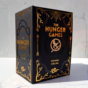 Puede incluir: Un libro de tapa dura negro titulado The Hunger Games de Suzanne Collins con detalles en lámina dorada. La portada presenta un emblema central de sinsajo, bordes de enredaderas ornamentados, flechas y un diseño de sol. El lomo muestra el título.