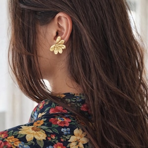 Puede incluir: Un pendiente de botón floral de color dorado se lleva en la oreja de una persona con cabello largo y castaño oscuro. La persona lleva una blusa con estampado floral azul oscuro con flores rojas, amarillas y azules.