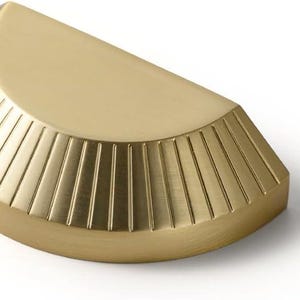 Peut inclure: Une poignée de meuble semi-circulaire avec une finition dorée brossée. La quincaillerie présente une surface supérieure plate et un bord incurvé orné de rainures verticales en creux qui rayonnent depuis le centre du bord droit.