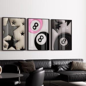 May include: Three framed art prints with black frames. The prints feature abstract designs: stars, a pink and black eight ball, and a black eight ball. The artwork hangs above a black leather sectional sofa.