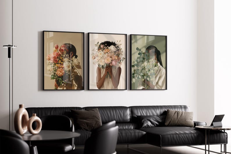 May include: Three framed art prints featuring figures with floral arrangements obscuring their faces. The prints have black frames and are hung above a black leather sectional sofa. The color palette includes beige, pink, and green.