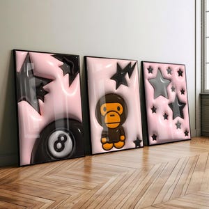 Op de afbeelding: Drie ingelijste wandkunstprints met een glanzende lichtroze achtergrond met 3D-effect. De eerste print toont een zwarte achtbal en zwarte sterren. De tweede print toont een bruin cartoon-aapfiguurtje en een zwarte ster. De derde print bevat meerdere grijze en zwarte sterren.