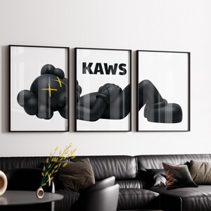 Puede incluir: Un conjunto de tres cuadros de pared que muestra a un personaje negro con ojos amarillos en forma de X tumbado a trav&eacute;s de tres impresiones enmarcadas. El panel central muestra el texto KAWS en letras may&uacute;sculas negras.