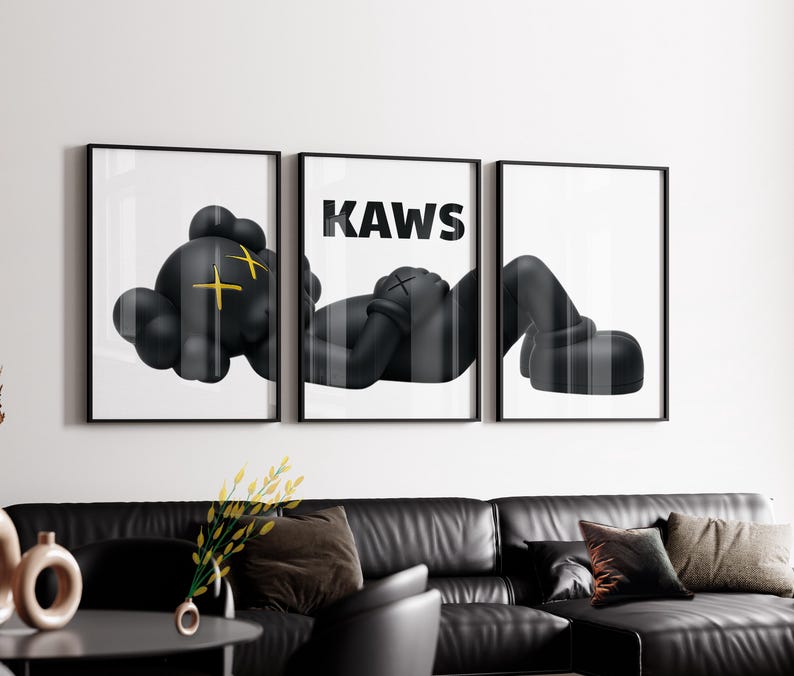 Puede incluir: Un conjunto de tres cuadros de pared que muestra a un personaje negro con ojos amarillos en forma de X tumbado a trav&eacute;s de tres impresiones enmarcadas. El panel central muestra el texto KAWS en letras may&uacute;sculas negras.
