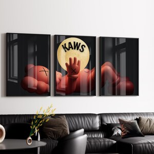 Puede incluir: Un conjunto de tres cuadros de pared que muestra a un personaje de dibujos animados rojo con ojos en forma de X acostado. El panel central muestra al personaje alcanzando una gran luna amarilla con el texto negro KAWS impreso arriba.