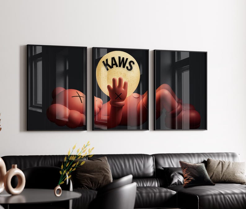 Puede incluir: Un conjunto de tres cuadros de pared que muestra a un personaje de dibujos animados rojo con ojos en forma de X acostado. El panel central muestra al personaje alcanzando una gran luna amarilla con el texto negro KAWS impreso arriba.