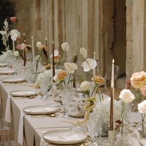 May include: A long table set for a special event, featuring a beige tablecloth, white plates, gold cutlery, and clear glassware. The centerpiece includes floral arrangements with white and peach roses, anthuriums, and tall brown candles.