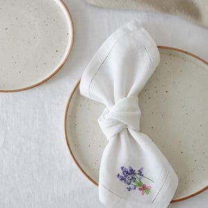 Puede incluir: Servilleta de lino blanco con bordado de lavanda y lazo rosa, anudada sobre un plato de cer&aacute;mica moteada con borde marr&oacute;n. La servilleta tiene un borde cosido. Un segundo plato es visible en el fondo, junto con un pa&ntilde;o de lino.