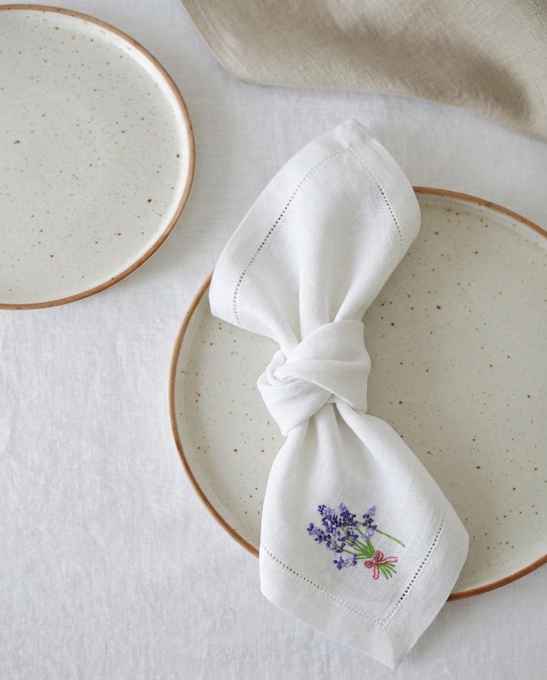 Puede incluir: Servilleta de lino blanco con bordado de lavanda y lazo rosa, anudada sobre un plato de cer&aacute;mica moteada con borde marr&oacute;n. La servilleta tiene un borde cosido. Un segundo plato es visible en el fondo, junto con un pa&ntilde;o de lino.