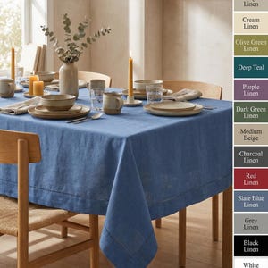 Può includere: Un tavolo da pranzo apparecchiato con una tovaglia di lino blu. Piatti, ciotole e tazze beige sono disposti sul tavolo. Candele gialle e un vaso con verde aggiungono al decoro. Sedie in legno circondano il tavolo.