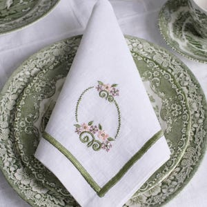 Peut inclure: Serviette en lin blanc avec une bordure brodée verte et un motif floral. La serviette est pliée et placée sur une assiette à motifs verts et blancs, faisant partie d'un service à thé. La broderie florale présente des accents roses et verts.