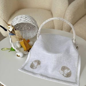 May include: A white Easter basket with a woven handle and a white linen cloth. The cloth is embroidered with two egg designs and a gray border. A smaller white basket with Easter decorations sits behind it.