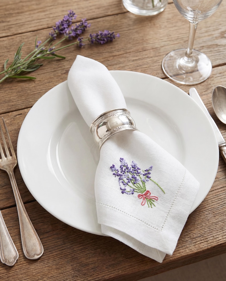 Puede incluir: Una servilleta de lino blanco, bordada con lavanda, se sujeta con un servilletero plateado sobre un plato blanco. Una ramita de lavanda y cubiertos est&aacute;n sobre una mesa de madera. La servilleta presenta un ramo de lavanda morada atado con una cinta rosa.