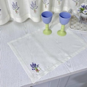 Puede incluir: Un mantel individual de lino blanco con un dise&ntilde;o bordado de lavanda. Dos copas azules y verdes est&aacute;n sobre el mantel. Cortinas bordadas a juego y una peque&ntilde;a bolsa en el fondo.