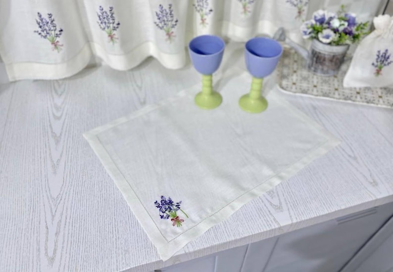 Puede incluir: Un mantel individual de lino blanco con un dise&ntilde;o bordado de lavanda. Dos copas azules y verdes est&aacute;n sobre el mantel. Cortinas bordadas a juego y una peque&ntilde;a bolsa en el fondo.