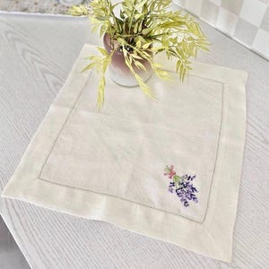 Puede incluir: Mantel individual de lino color crema con un delicado ramo de lavanda bordado y atado con una cinta rosa. El mantel individual tiene un borde cosido. Un jarr&oacute;n con vegetaci&oacute;n est&aacute; en el fondo, sobre una superficie de madera clara.