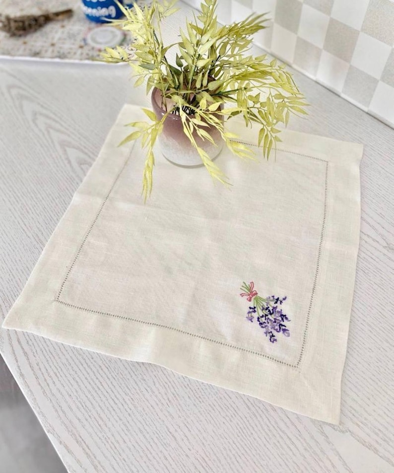 Puede incluir: Mantel individual de lino color crema con un delicado ramo de lavanda bordado y atado con una cinta rosa. El mantel individual tiene un borde cosido. Un jarr&oacute;n con vegetaci&oacute;n est&aacute; en el fondo, sobre una superficie de madera clara.