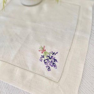 Puede incluir: Mantel individual de lino color crema con un delicado ramo de lavanda bordado atado con una cinta rosa. El mantel individual tiene un borde cosido y est&aacute; colocado sobre una superficie de color claro. El bordado presenta tonos de morado y verde.