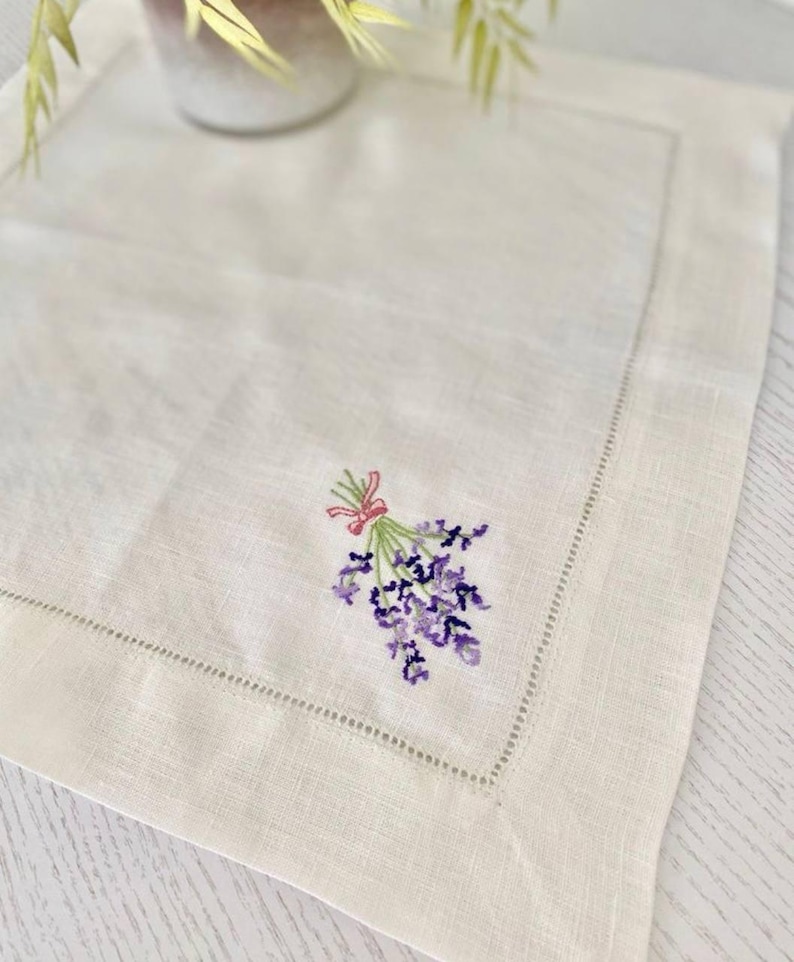 Puede incluir: Mantel individual de lino color crema con un delicado ramo de lavanda bordado atado con una cinta rosa. El mantel individual tiene un borde cosido y est&aacute; colocado sobre una superficie de color claro. El bordado presenta tonos de morado y verde.