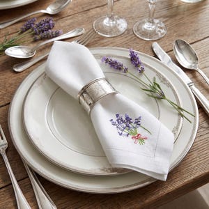 Puede incluir: Una elegante mesa con platos blancos, cubiertos y copas de cristal. Una servilleta de lino blanca, bordada con lavanda, est&aacute; sujeta por un servilletero plateado. Ramas de lavanda a&ntilde;aden un toque natural a la disposici&oacute;n.
