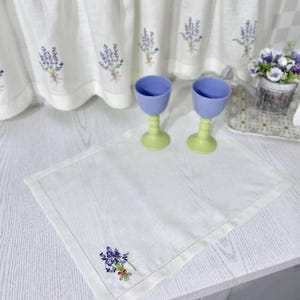Puede incluir: Mantel individual de lino blanco con un dise&ntilde;o de bordado de lavanda. Dos copas azules y verdes est&aacute;n sobre la mesa. Cortinas a juego con bordados de lavanda en el fondo. Tambi&eacute;n se ve una peque&ntilde;a maceta con flores.