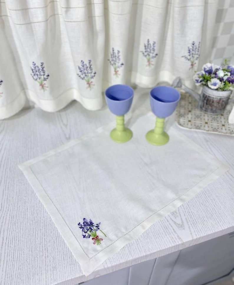 Puede incluir: Mantel individual de lino blanco con un dise&ntilde;o de bordado de lavanda. Dos copas azules y verdes est&aacute;n sobre la mesa. Cortinas a juego con bordados de lavanda en el fondo. Tambi&eacute;n se ve una peque&ntilde;a maceta con flores.