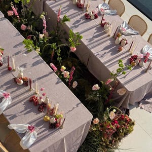 Peut inclure: Vue aérienne de deux tables dressées pour un événement. Les nappes mauves sont ornées de compositions florales, de raisins et de bougies. Des chaises en osier entourent les tables, et la scène est baignée de lumière naturelle.