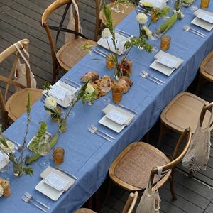 May include: An overhead shot of a beautifully set table for an outdoor event. A long, light blue tablecloth is adorned with white plates, silverware, and amber-colored glasses. Floral arrangements and bread add to the elegant ambiance. Wooden chairs surround the table.