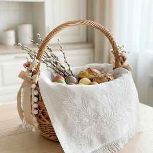 Op de afbeelding: Een geweven paasmand met een handvat, gevuld met versierde eieren, gebakken goederen en katjeswilgtakken. Een witte geborduurde linnen doek met franjes en bloemendetails bedekt de inhoud. De mand is versierd met linten en pompons.
